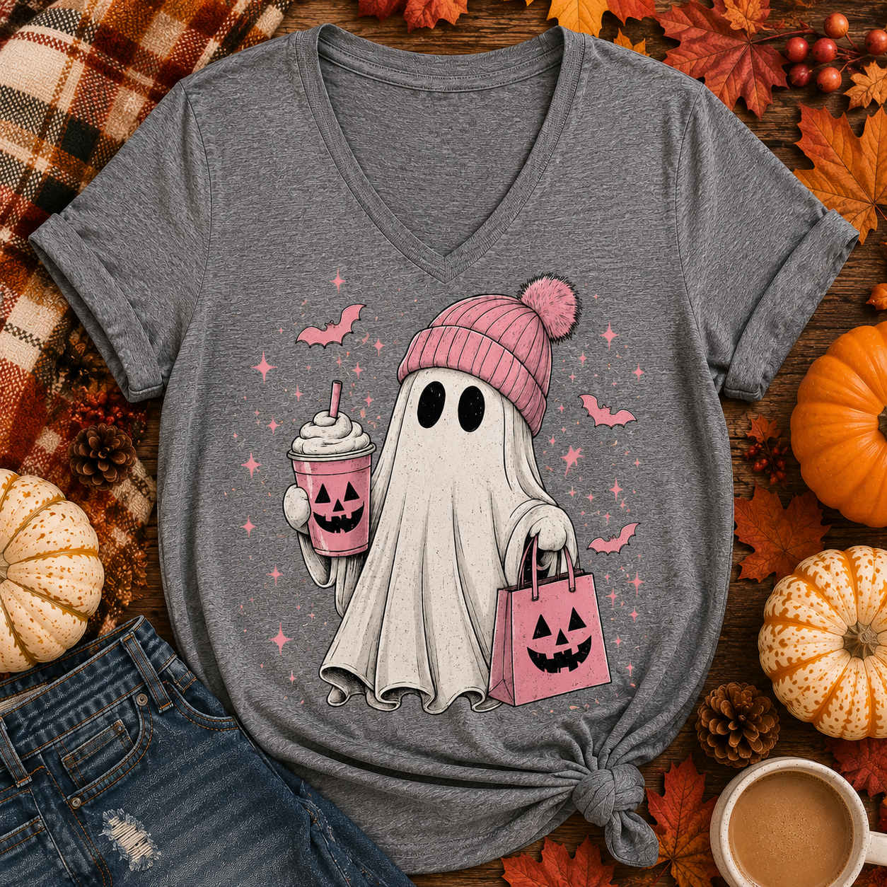 A heather gray V-neck t-shirt with a graphic of a cute cartoon ghost wearing a pink knit beanie and holding a jack-o'-lantern drink and shopping bag. The shirt is styled in a flat-lay arrangement with autumn props including pumpkins, fall leaves, pinecones, a plaid blanket, and a coffee cup.