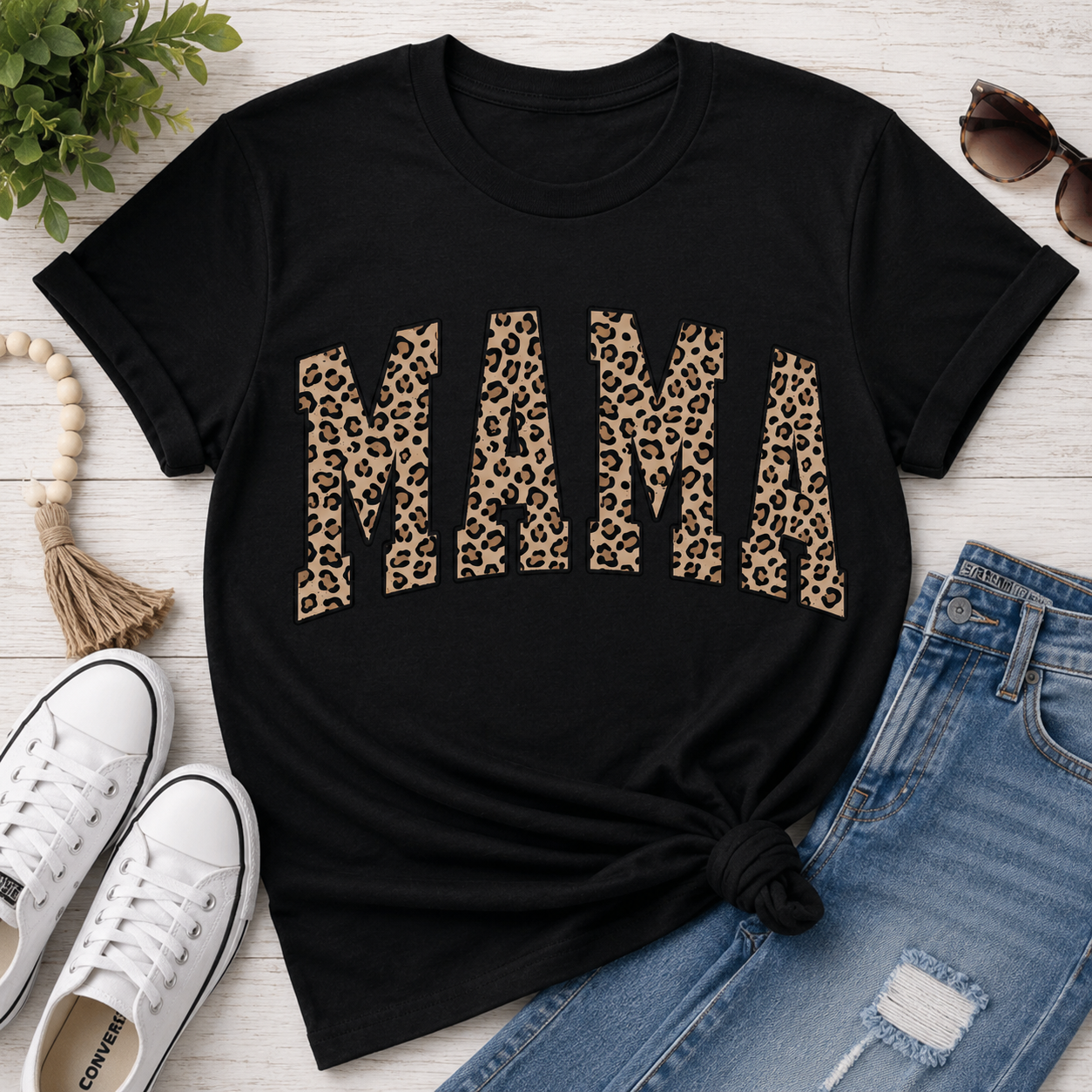Black graphic t-shirt featuring bold leopard print "MAMA" lettering, styled with distressed blue jeans, white sneakers, sunglasses, and neutral accessories for a casual everyday mom outfit.